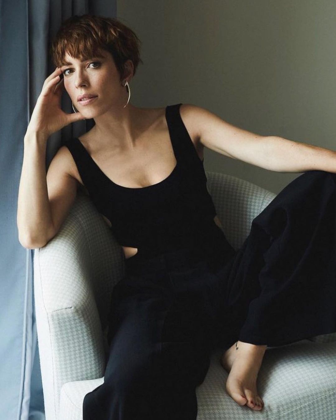 Attractive woman with short auburn hair in a black top and wide leg pants sits in a grey chair in an indoor setting by a window.