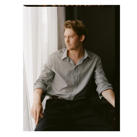 A man wearing a striped button-down shirt and dark trousers seated by a window, photographed by Victoria Stevens.