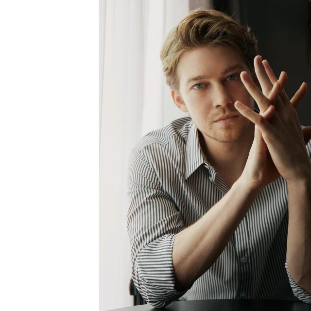 Portrait of a blond adult male in a striped shirt, clasping hands together near his face, with soft window light.