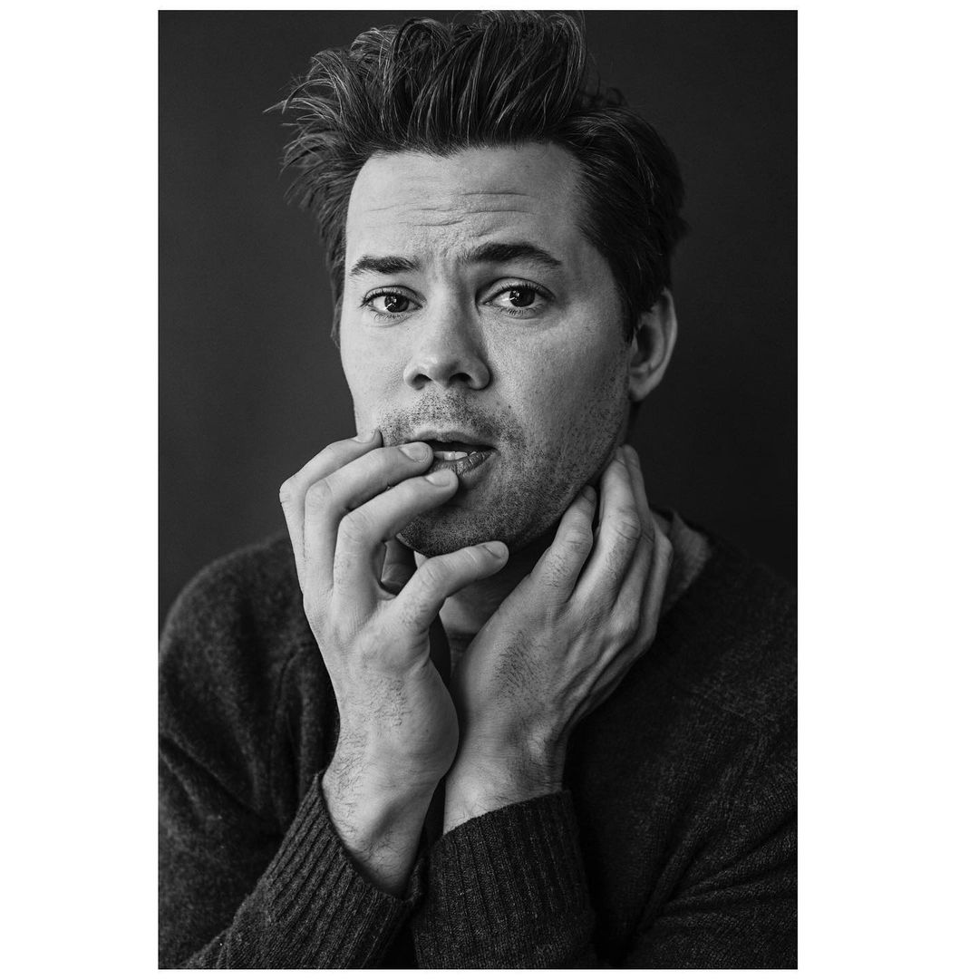 Black and white portrait of an adult male with dark hair, wearing a sweater, looking at the camera, and touching his face with his hands.