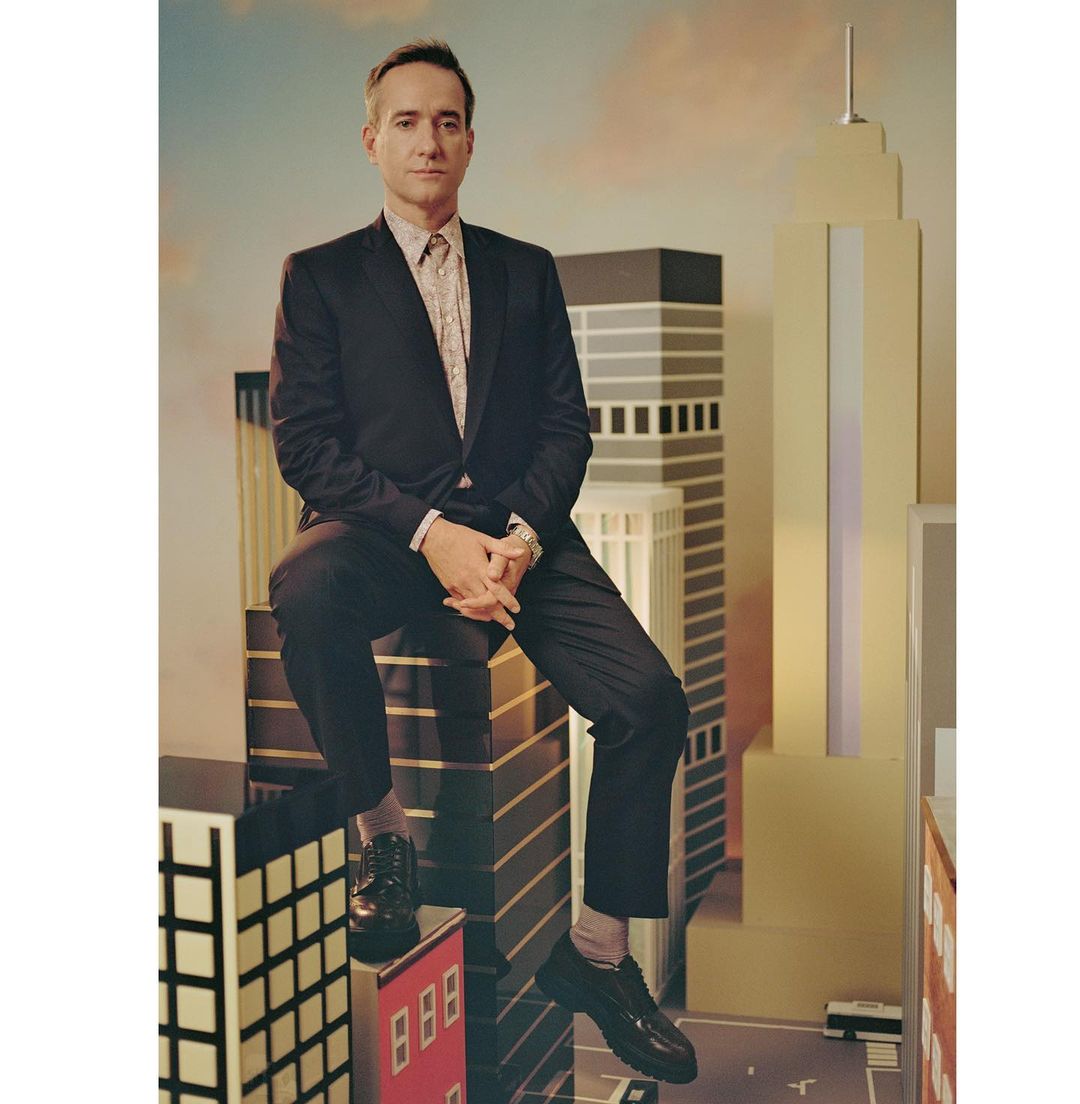 Formally dressed adult man sitting atop faux skyscraper with a neutral facial expression, wearing a suit and patterned shirt.