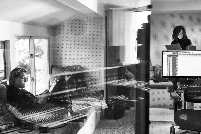 Two people inside a recording studio, with a man seated and using a mixing console, and a woman using a laptop in the background.