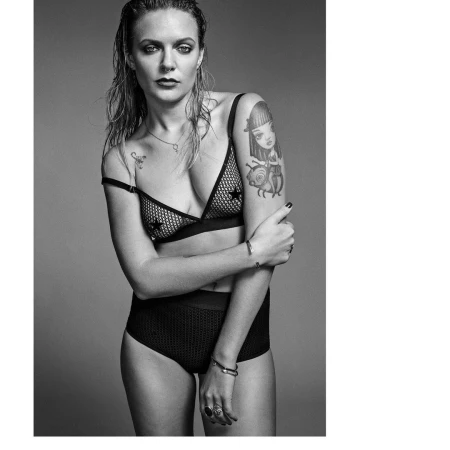 Portrait of a woman with wet hair wearing a black mesh bra and high-waisted briefs against a grey studio backdrop.
