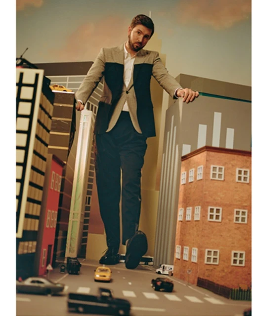 Adult male in a two-tone jacket stands in a miniature city street with toy cars and tall buildings, shot at a low angle.