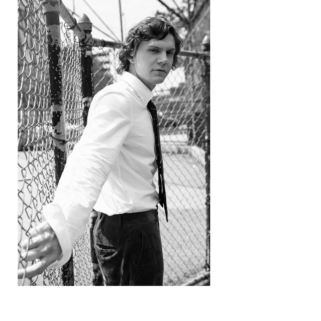 Black and white portrait of Evan Peters wearing a white button-down shirt and tie, leaning against a chain-link fence.