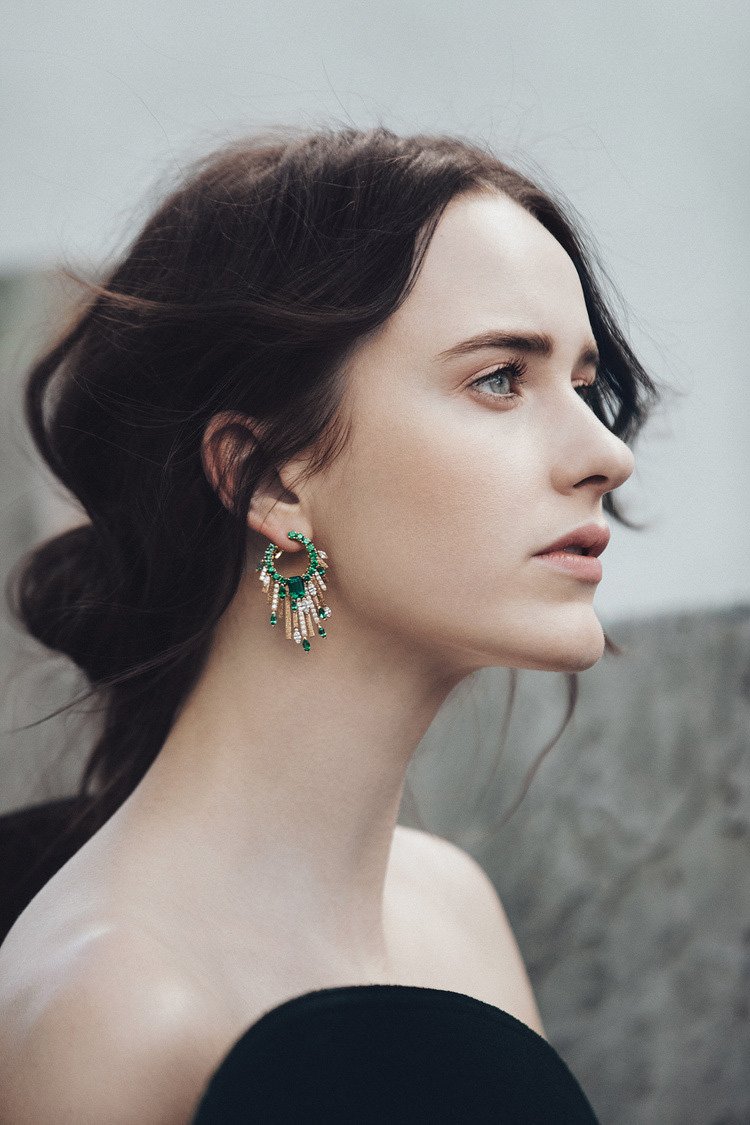 Elegant woman in profile wearing dangling emerald and crystal earrings, with dark hair pulled back and blurred neutral background.