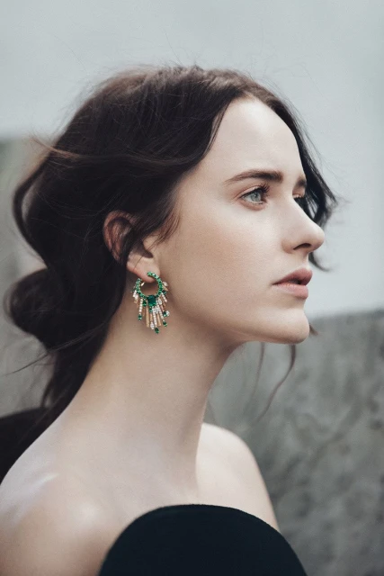 Elegant woman in profile wearing dangling emerald and crystal earrings, with dark hair pulled back and blurred neutral background.