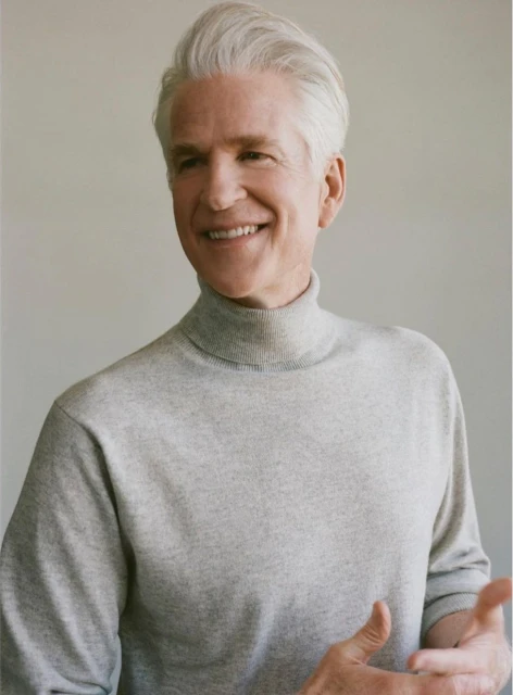 Portrait of smiling adult male with grey hair wearing grey turtleneck sweater indoors against a plain background in soft lighting.