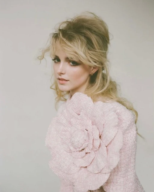 Portrait of a blonde woman in a pink dress with a large flower detail against a neutral background in a studio fashion photograph.
