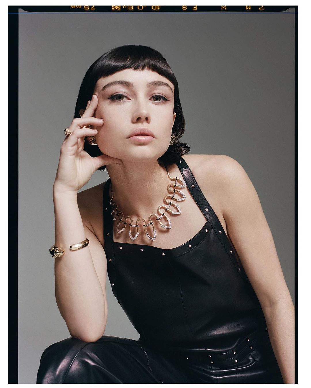 A young woman with dark hair and bangs wears a statement necklace and leather outfit against a grey backdrop in a studio fashion shot.