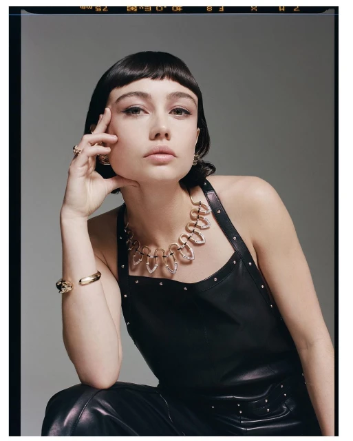 A young woman with dark hair and bangs wears a statement necklace and leather outfit against a grey backdrop in a studio fashion shot.