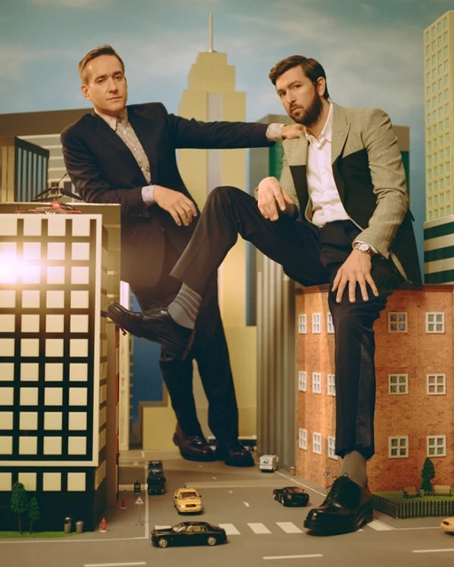 Two men in suits pose playfully on miniature city buildings, creating a giant effect against a backdrop of toy cars and skyscrapers.