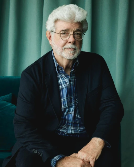 Portrait of George Lucas wearing glasses and a black jacket, seated in front of a teal-colored textured wall and teal velvet couch.