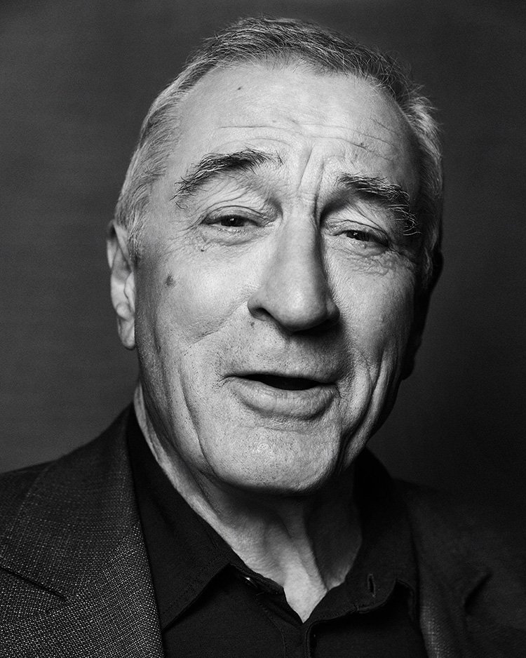 Monochrome portrait of Robert De Niro, an elderly man with short gray hair wearing a dark collared shirt and a grey jacket indoors.