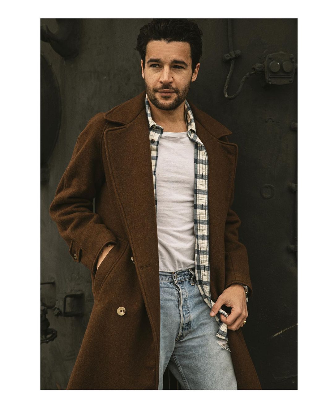 Portrait of a stylish adult male with dark hair wearing a brown overcoat, plaid shirt, and ripped jeans standing against a dark background.