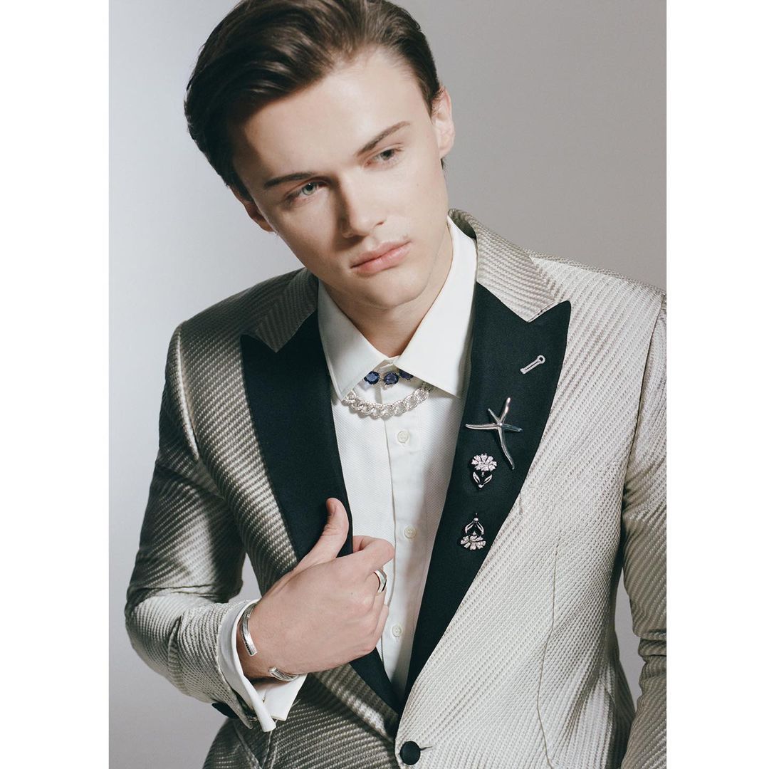 Young man in a textured suit with jewelry details. He wears a pearl necklace and decorative pins on his suit jacket in a studio setting.