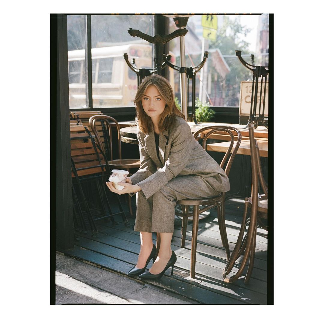 A woman in a grey suit sits on a chair outside a cafe, holding a white object, with sunlight streaming onto a wooden floor.