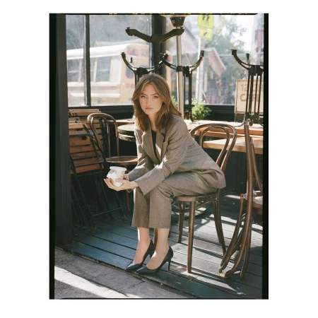 A woman in a grey suit sits on a chair outside a cafe, holding a white object, with sunlight streaming onto a wooden floor.