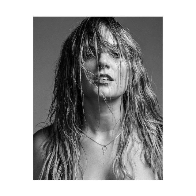 Black and white portrait of a woman with wet blonde hair, a nose ring, and a Venus symbol necklace against a grey backdrop.