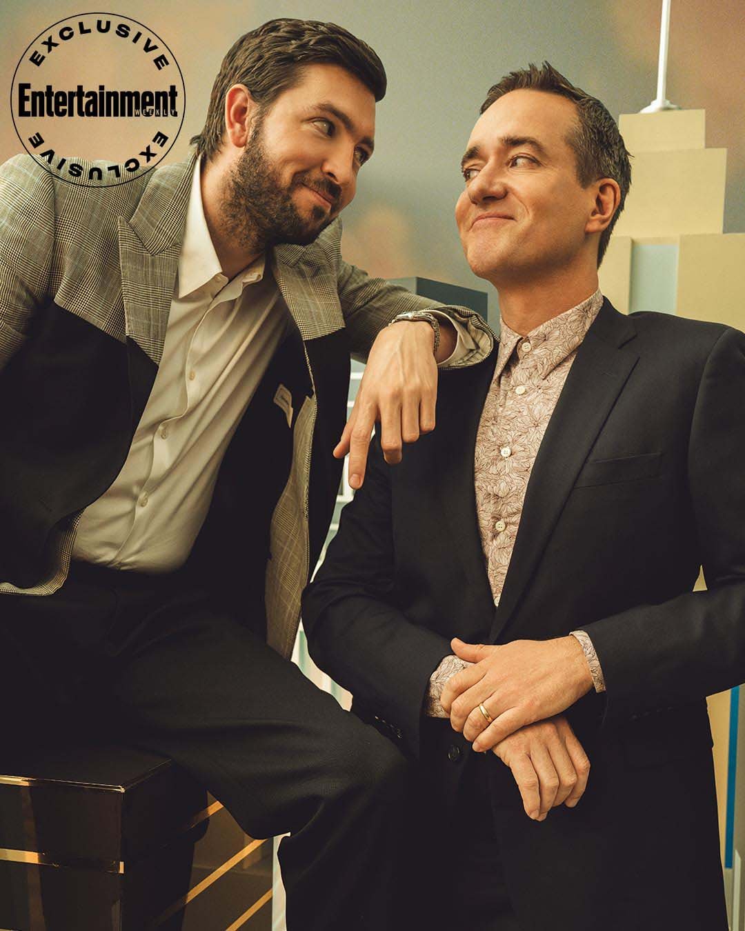 Two men pose in formal attire against a retro backdrop. One man is leaning while looking at the other who has his hands clasped.