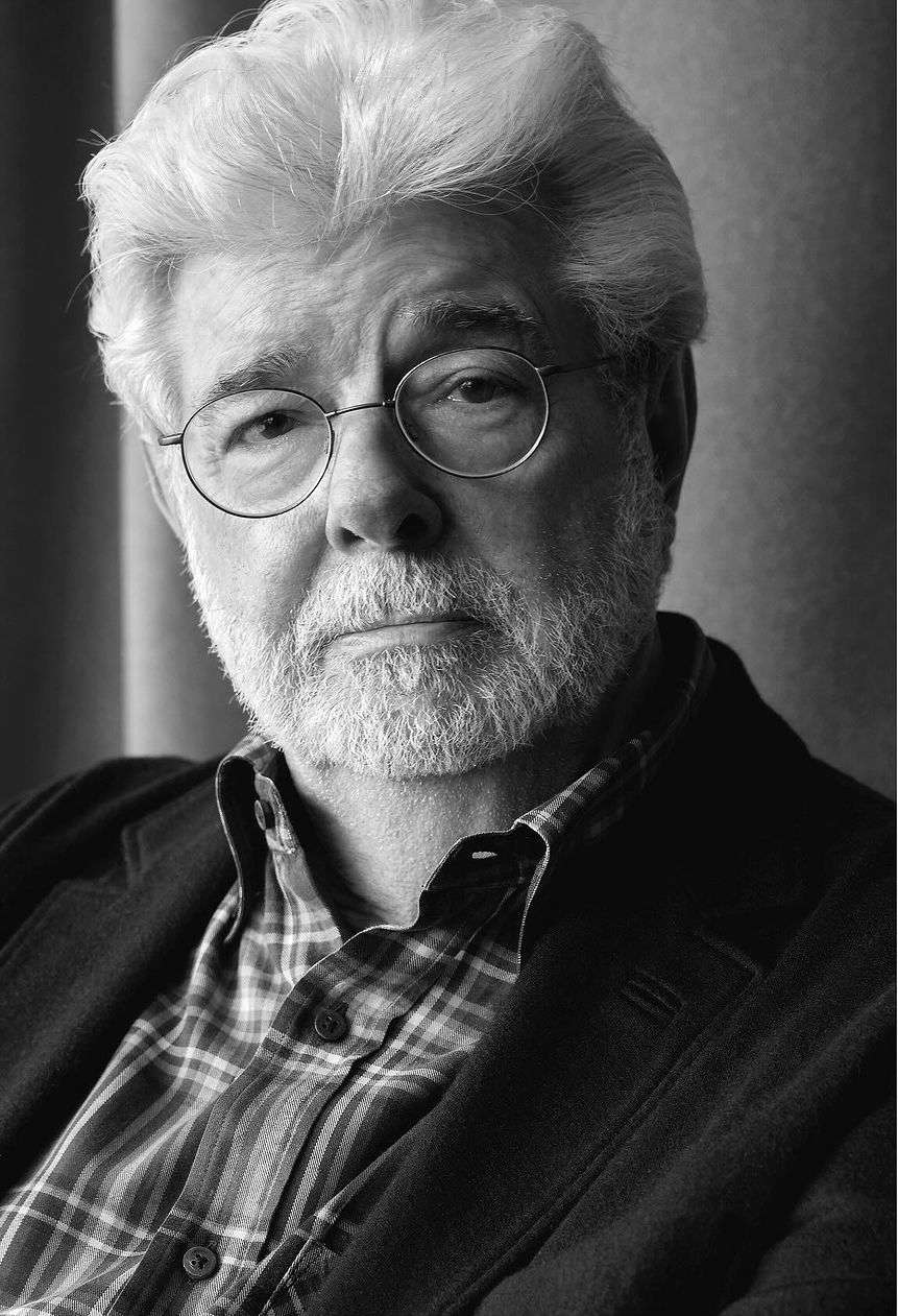Monochrome portrait of an older man with glasses, gray hair, and a beard, wearing a patterned shirt and blazer in a studio setting.