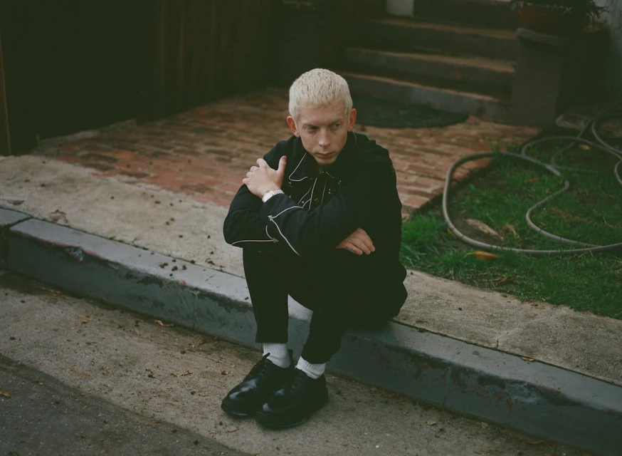 Young adult with platinum blonde hair sits on a concrete curb outside a brick home, wearing a black jacket and shoes on a cloudy day.