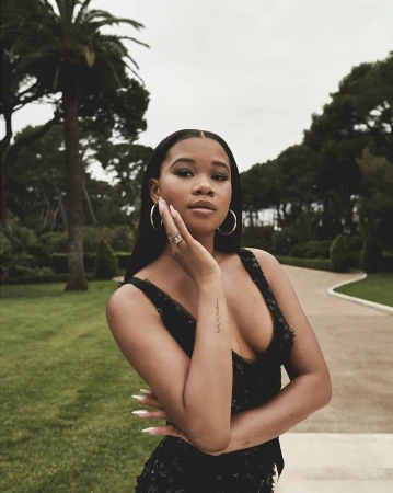 Portrait of a young black woman in a black dress with gold hoop earrings outdoors against a backdrop of trees and a paved path
