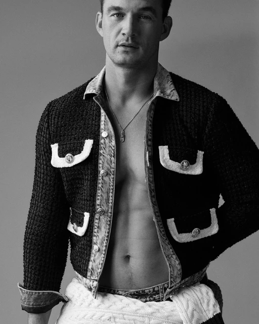 Portrait of a handsome adult male wearing a black tweed jacket with denim details and a silver necklace, standing against a grey backdrop.