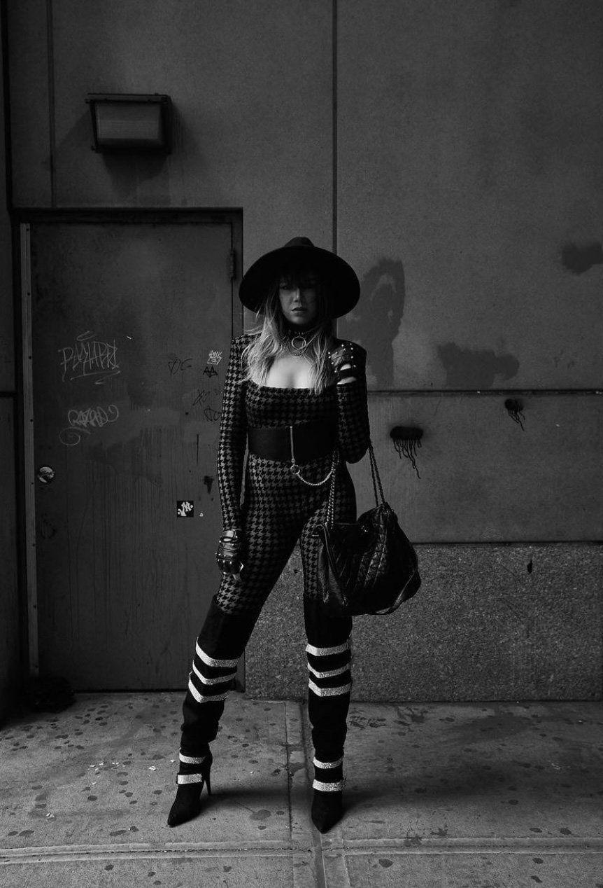 A woman poses in a houndstooth jumpsuit, a wide-brimmed hat, and high-heeled boots on a city street with a textured wall and door.