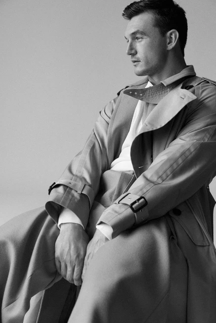 Black-and-white portrait of a stylish adult male wearing a trench coat and sweater in a studio setting looking off to the side.