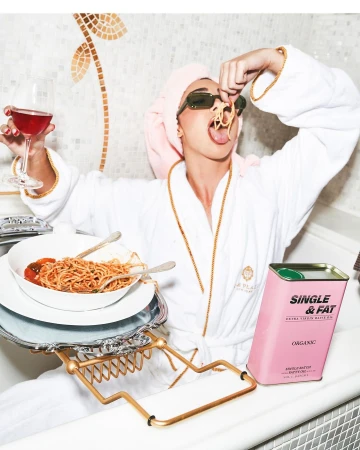 A woman in a bathrobe and sunglasses sits in a bathtub eating spaghetti and drinking wine, with a can of olive oil nearby.