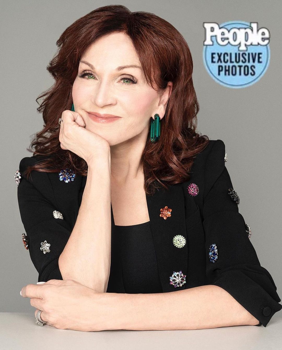 Headshot of a mature woman with auburn hair wearing a black blazer adorned with decorative pins, posing against a grey background.