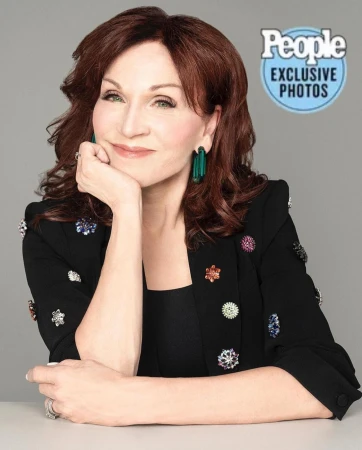 Headshot of a mature woman with auburn hair wearing a black blazer adorned with decorative pins, posing against a grey background.
