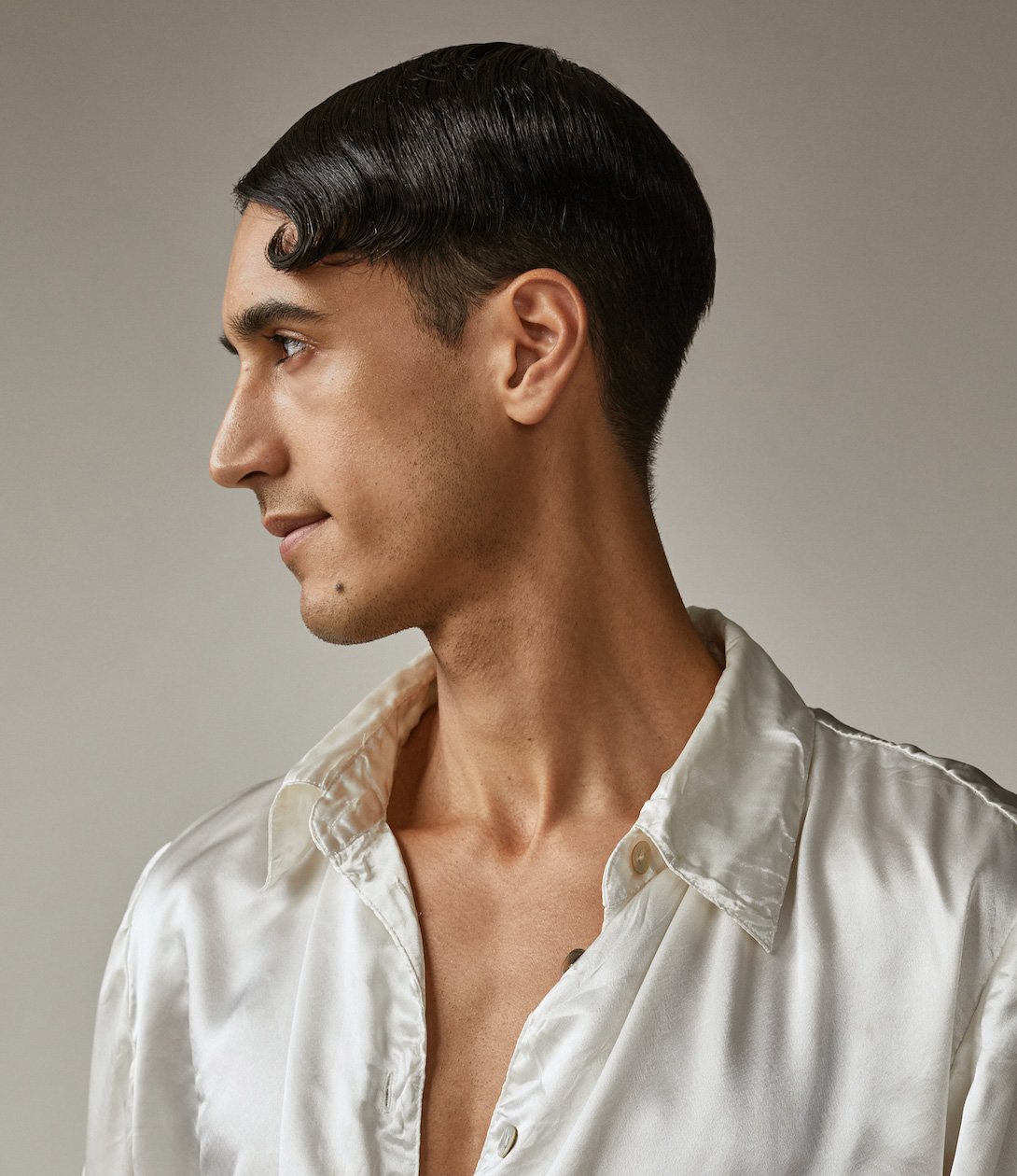 Profile view of a young adult male with dark wavy hair, wearing an unbuttoned white satin shirt against a neutral background.