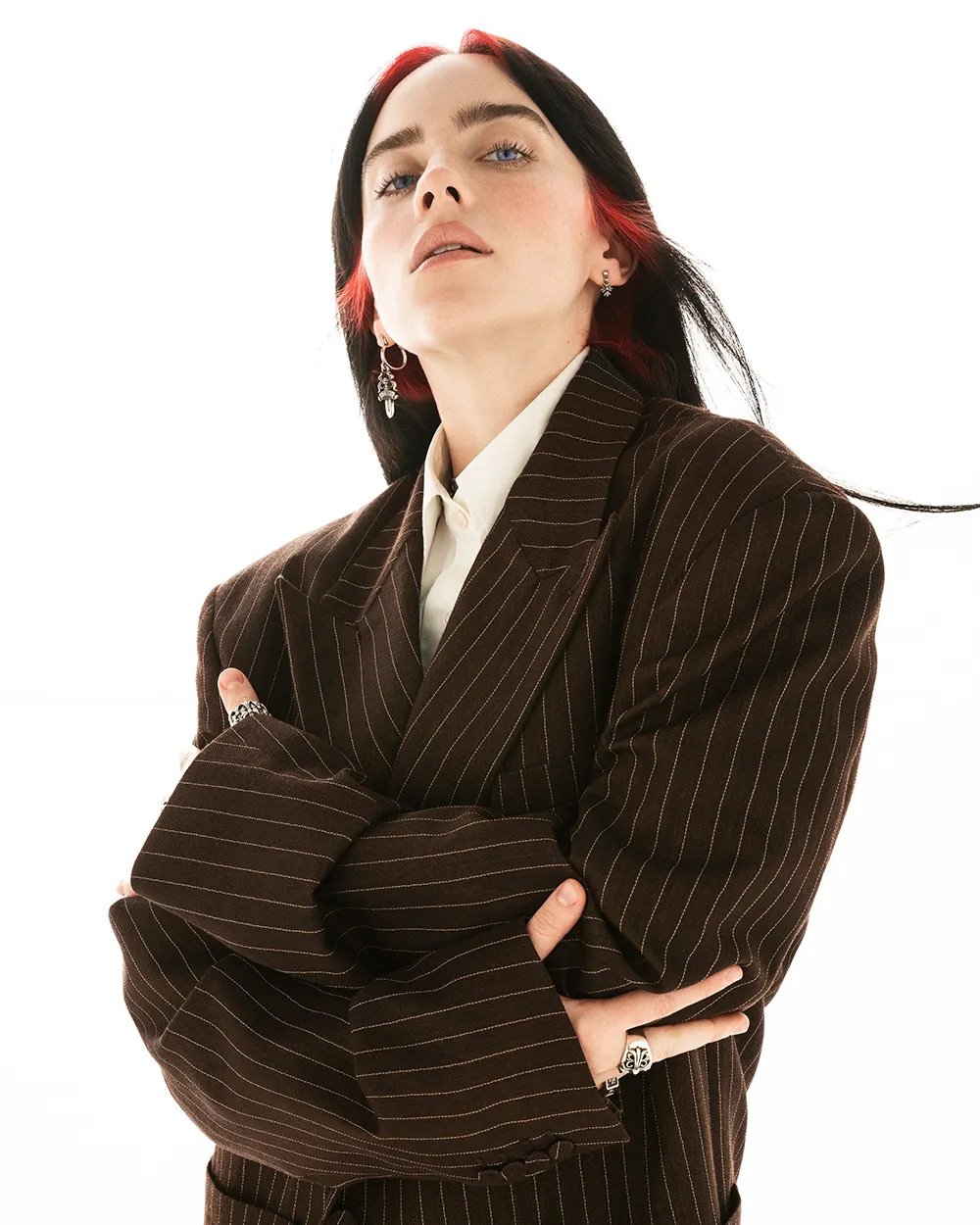Portrait of Billie Eilish in a pinstripe suit, crossing her arms against a bright white backdrop, accentuated by her dark hair with red highlights.