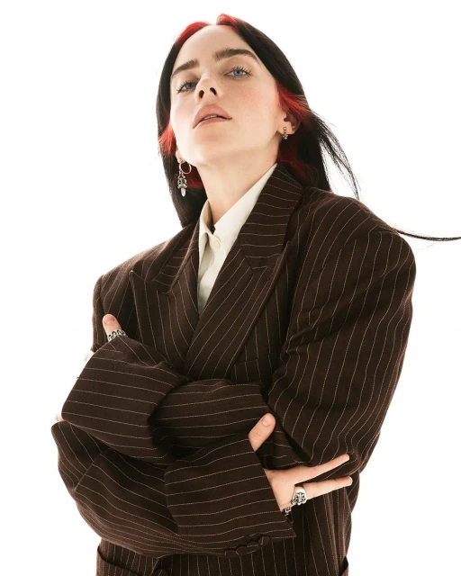 Portrait of Billie Eilish in a pinstripe suit, crossing her arms against a bright white backdrop, accentuated by her dark hair with red highlights.
