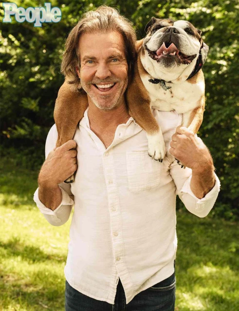 Adult male smiles while carrying a bulldog on his shoulders outdoors in a medium shot with bright lighting and green background.