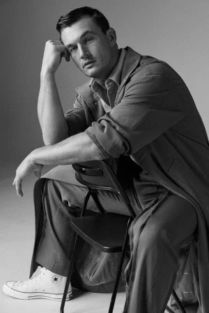 Monochrome portrait of a man sitting backward on a folding chair wearing a long coat and sneakers against a seamless background