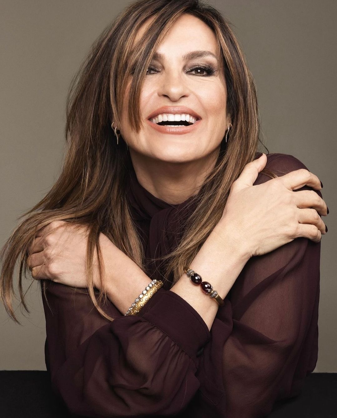 A medium shot captures a smiling adult woman with long brown hair, wearing a sheer brown top and bracelets, posing indoors against a neutral backdrop.