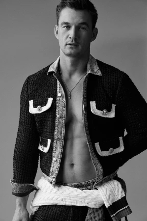 Portrait of a man wearing an open textured jacket and layered knitwear against a solid background.
