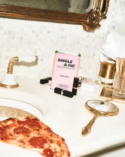 Bathroom sink setup: slice of pizza, pink can labeled 'Single & Fat,' lipsticks, gold mirror, gold fixtures, mosaic tile backsplash.