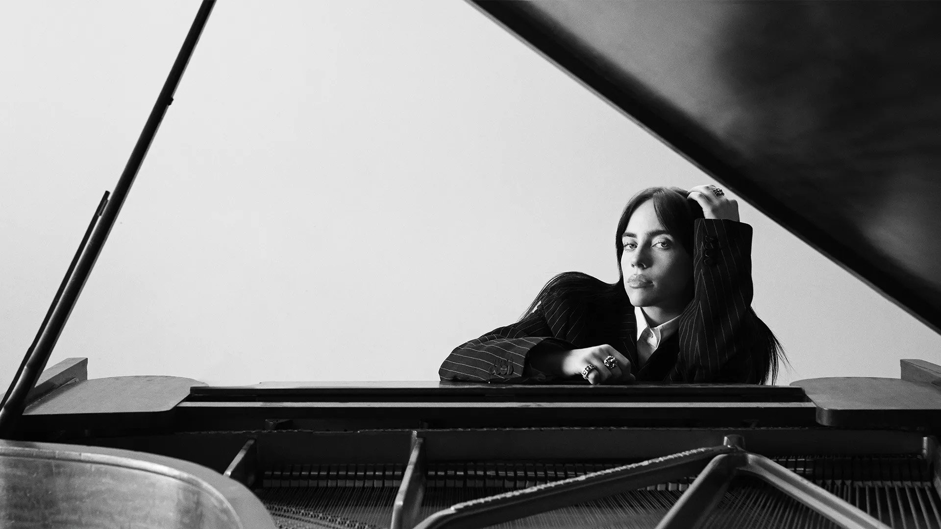 Black and white portrait of a young adult woman with dark hair leaning on a grand piano, wearing a suit jacket with rings on her fingers.