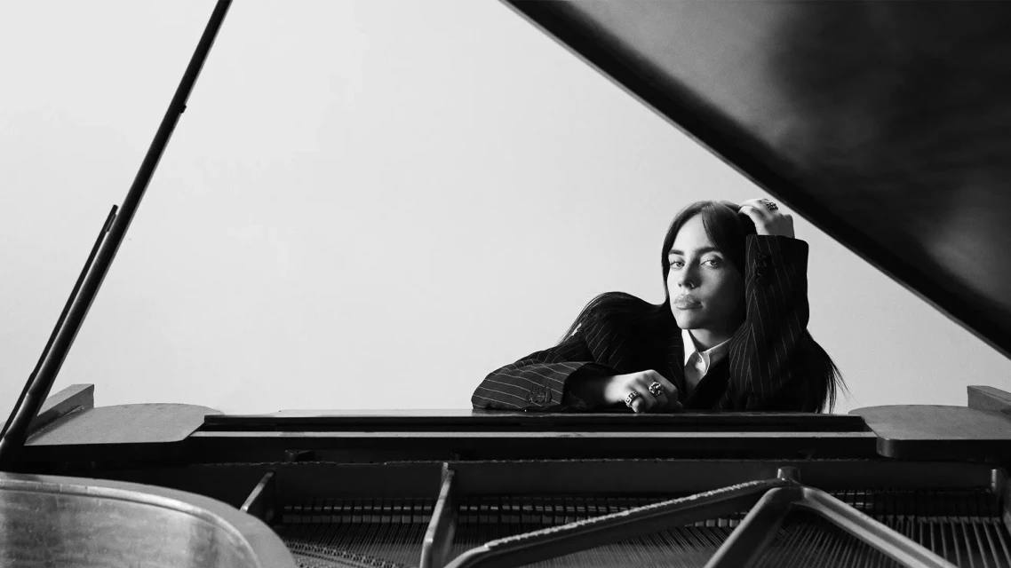 Black and white portrait of a young adult woman with dark hair leaning on a grand piano, wearing a suit jacket with rings on her fingers.