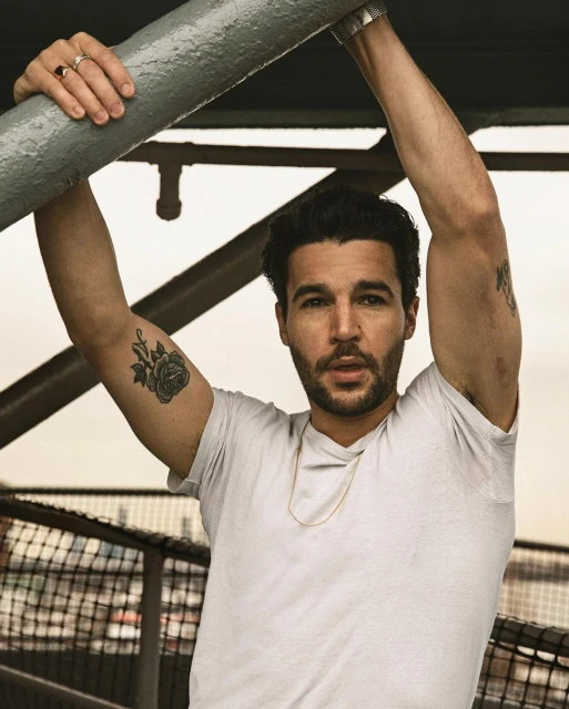 Adult male with arm tattoos poses outdoors, leaning on a gray metal pipe, wearing a white t-shirt, gold chain, and rings; close-up.