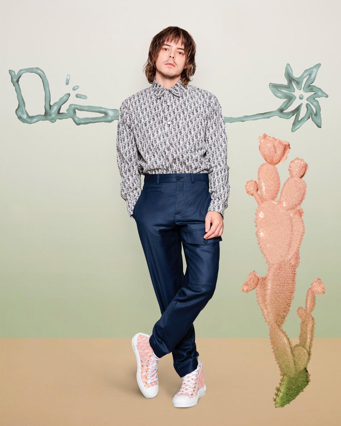 Adult male model in patterned shirt and dark pants, posing in a studio with cactus and painted designs against a two-tone backdrop.