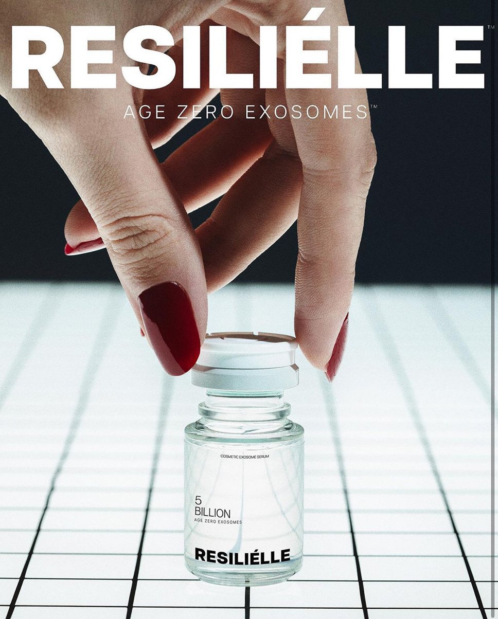 Close-up of an adult female hand holding a clear bottle of Resiliélle skin serum with red nail polish and a white grid background.