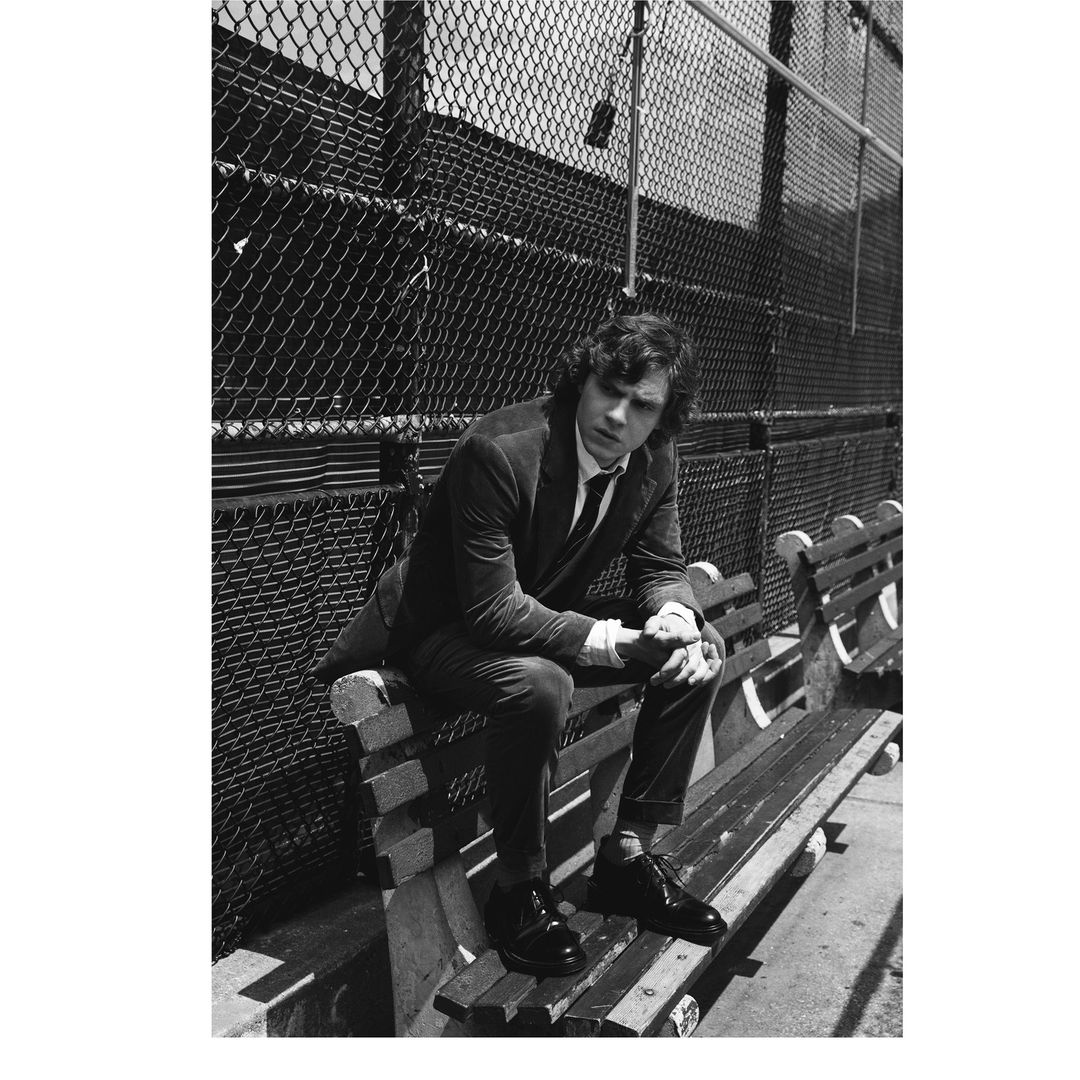 Man in a dark suit sitting on a wooden bench outdoors against a chain link fence in a black and white portrait.