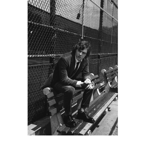 Man in a dark suit sitting on a wooden bench outdoors against a chain link fence in a black and white portrait.