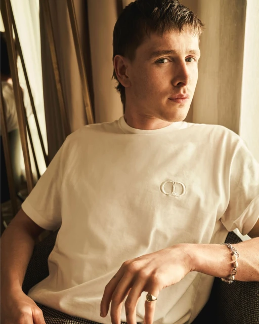 Portrait of a young adult male wearing a white t-shirt with a CD logo, sitting in an indoor setting, with soft lighting and a shallow depth of field.
