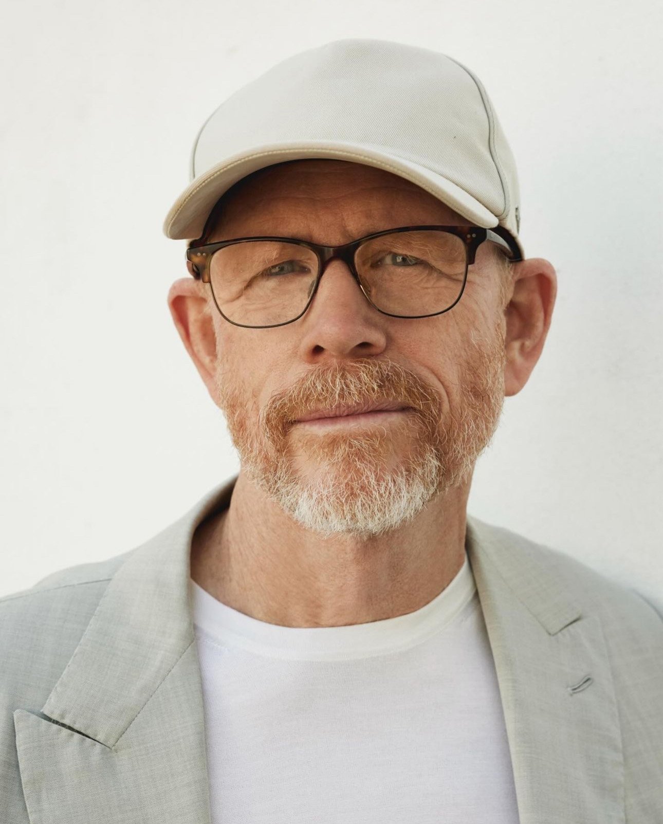 Portrait of Ron Howard wearing glasses, a white baseball cap, white t-shirt, and light grey blazer against a plain white background